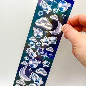 May include: A sheet of iridescent holographic stickers featuring a variety of celestial designs, including stars, moons, clouds, and shooting stars.