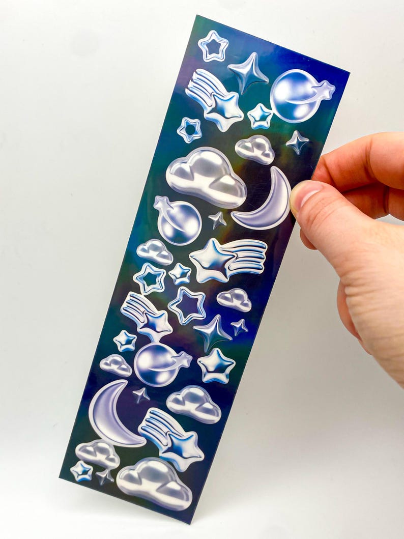 May include: A sheet of iridescent holographic stickers featuring a variety of celestial designs, including stars, moons, clouds, and shooting stars.