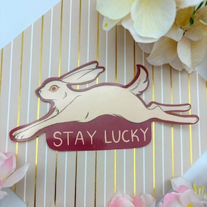 May include: A sticker featuring a running rabbit in cream and burgundy. The rabbit is leaping across a burgundy banner that reads "STAY LUCKY" in white text. The background has a striped pattern with gold accents.