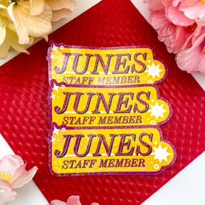 May include: Three yellow and purple "JUNES STAFF MEMBER" name tags with star accents. The name tags are stacked on a textured red surface, with pink and yellow flowers in the background.