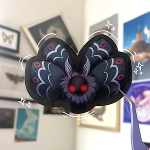 May include: A black and blue moth sticker with red eyes and white accents. The sticker is being held by tweezers and has a shiny finish.