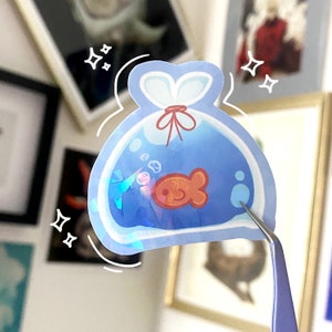 May include: A blue iridescent sticker of a goldfish in a plastic bag. The bag has a red ribbon tied around it. The sticker is on a white background.