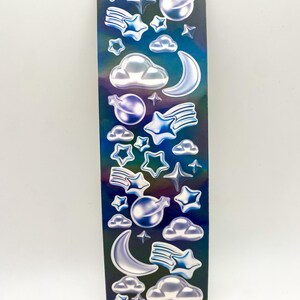May include: A sheet of iridescent blue and purple stickers featuring various celestial shapes, including stars, moons, clouds, and shooting stars. The stickers are 3D and have a glossy finish.