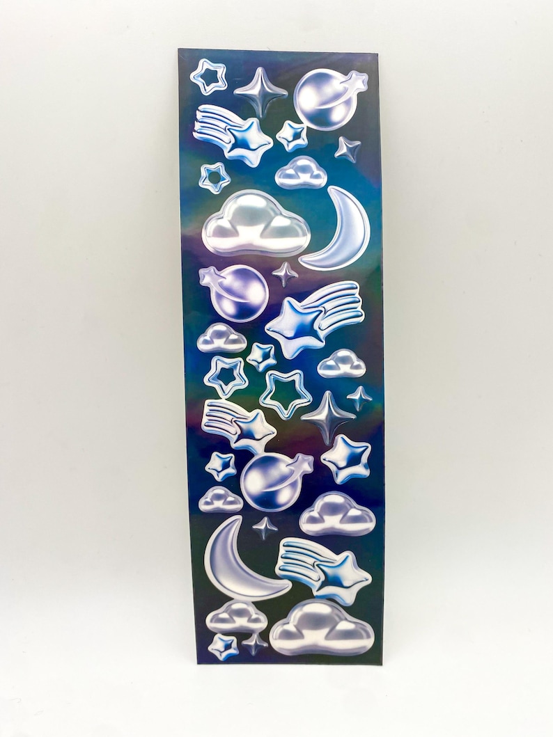 May include: A sheet of iridescent blue and purple stickers featuring various celestial shapes, including stars, moons, clouds, and shooting stars. The stickers are 3D and have a glossy finish.