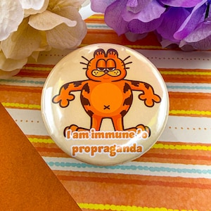 May include: A round button featuring a cartoon cat with orange fur and brown spots. The cat has a grumpy expression and is standing with its arms outstretched. The text "I am immune propaganda" is printed on the button.