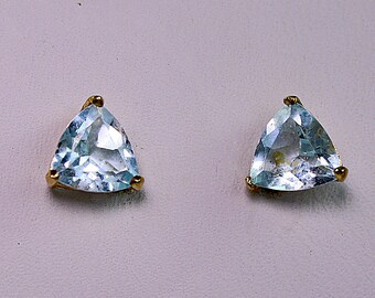 May include: A pair of stud earrings featuring light blue, triangular-cut gemstones set in gold-toned metal prongs. Each earring displays a faceted surface that reflects light, showcasing a clear, pale blue hue held securely by three small metal claws.