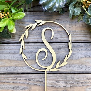Peut inclure: Un cake topper en bois doré avec la lettre "S" à l'intérieur d'une couronne circulaire de feuilles. Le topper est conçu pour les gâteaux et autres desserts de fête. Le fond est une surface en bois avec de la verdure.