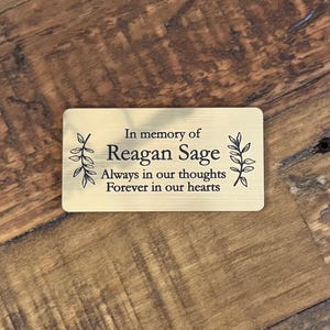 May include: A rectangular, gold-colored memorial plaque with the inscription "In memory of Reagan Sage. Always in our thoughts, Forever in our hearts." The plaque is decorated with leaf sprigs and rests on a wooden surface.