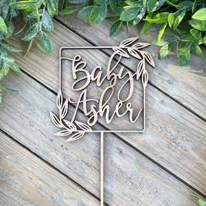 Puede incluir: Topper de pastel de madera con el texto "Baby Asher" en un marco cuadrado con acentos de hojas.