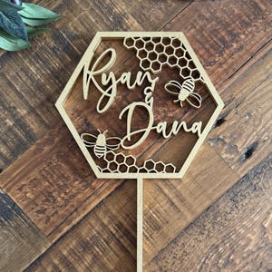 Puede incluir: Un adorno para pastel de madera dorada en forma hexagonal, con los nombres "Ryan & Dana" en escritura cursiva. El diseño incluye patrones de panal y dos ilustraciones de abejas, ideal para una boda o compromiso.