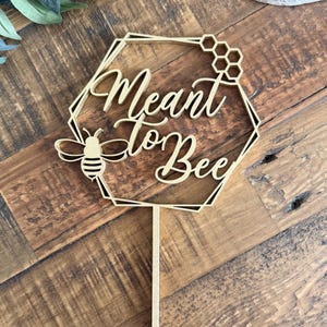 Puede incluir: Un adorno para pastel de madera con la frase "Meant to Bee" en una elegante escritura. El diseño incluye un marco hexagonal, un panal y una ilustración de abeja. El adorno es de color marrón claro y tiene un pincho largo y puntiagudo.