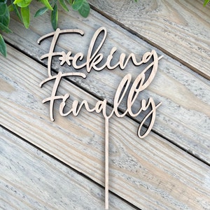 Pode incluir: Topper de bolo de madeira com o texto "F*cking Finally" em uma fonte cursiva.