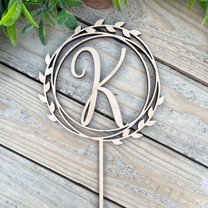 Peut inclure: Un cake topper en bois avec la lettre "K" au centre d'un cercle orné de feuilles. Le topper est doté d'une longue tige pointue. Fabriqué en bois clair, il est idéal pour décorer un gâteau de fête.