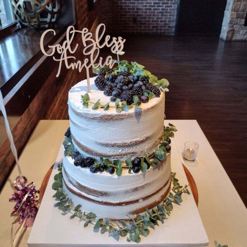 May include: A two-tiered naked cake decorated with fresh blueberries and greenery. The cake is topped with a wooden sign that reads "God Bless Amelia".