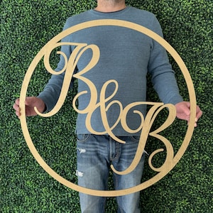 May include: A gold wooden circle with the letters "B & B" in a cursive font. The circle is being held by a person.
