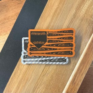 May include: A brown leather sticker with a baseball home plate and bats arranged in the shape of a British flag. The sticker is on a wooden surface.
