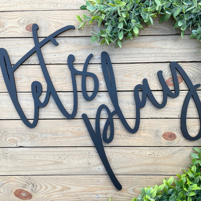 May include: Black metal sign with the words "Let's Play" written in a cursive font. The sign is hanging on a wooden wall.