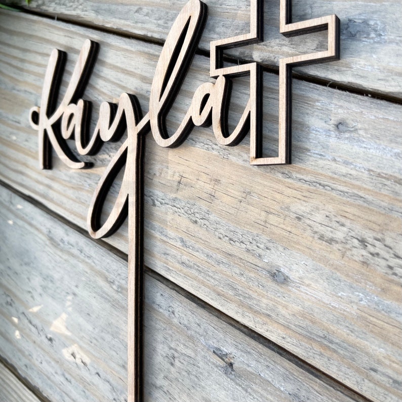 K&ouml;nnte beinhalten: Holzausschnitt des Namens "Kayla" mit einem Kreuz dahinter. Der Ausschnitt befindet sich auf einem hellbraunen Holzgrund.