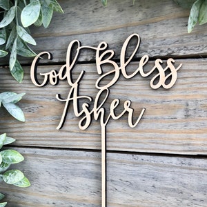 May include: Wooden cake topper with the text "God Bless Asher" in a cursive font. The topper is light brown and has a thin, black stick attached to the back.