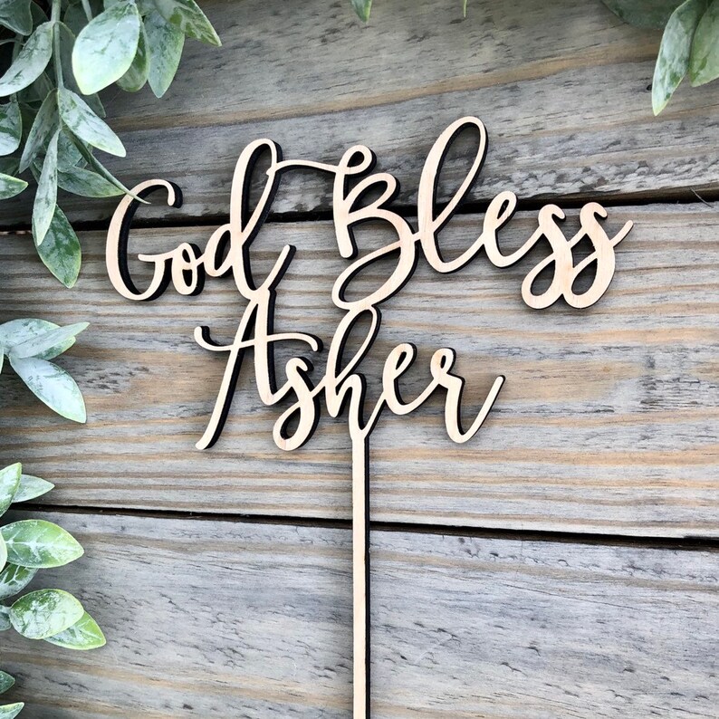 May include: Wooden cake topper with the text "God Bless Asher" in a cursive font. The topper is light brown and has a thin, black stick attached to the back.
