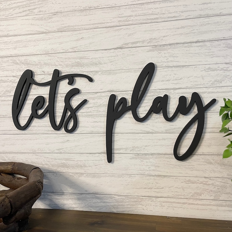 K&ouml;nnte beinhalten: Schwarzes Holzschild mit den Worten "Let's Play" in einer kursiv geschriebenen Schrift.