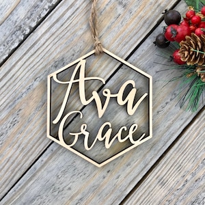 May include: A wooden hexagon ornament with the names "Ava Grace" written in cursive script. The ornament is hanging from a twine string and is on a wood background.