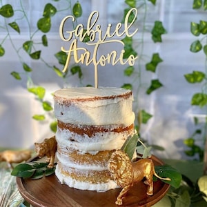 May include: A three-tiered cake with white frosting and a wooden cake topper that says "Gabriel Antonio". The cake is decorated with two gold lion figurines.