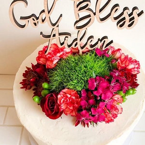 May include: A white cake with a wooden cake topper that says "God Bless Aiden" and is decorated with red and green flowers.