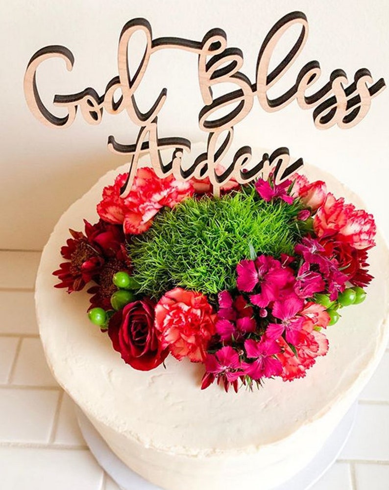 May include: A white cake with a wooden cake topper that says "God Bless Aiden" and is decorated with red and green flowers.