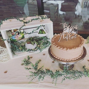 May include: A white wooden crate with greenery and a photo inside. A chocolate cake with a wooden sign that says "Becca & Aaron" is on a silver cake stand. The cake and crate are on a white tablecloth with greenery.