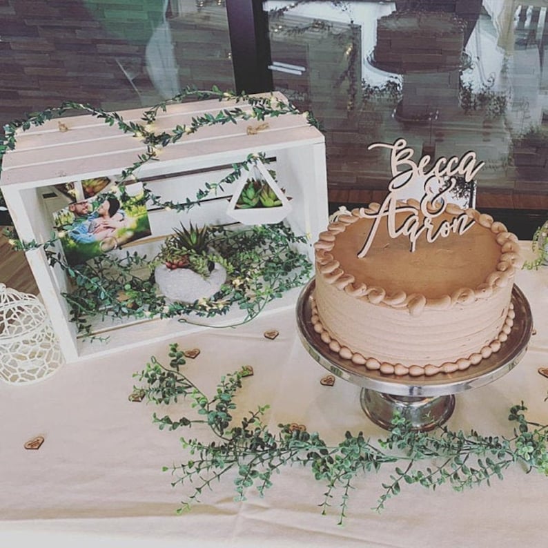 May include: A white wooden crate with greenery and a photo inside. A chocolate cake with a wooden sign that says "Becca & Aaron" is on a silver cake stand. The cake and crate are on a white tablecloth with greenery.