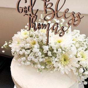 May include: A white cake with white flowers and a wooden cake topper that says "God Bless Thomas".