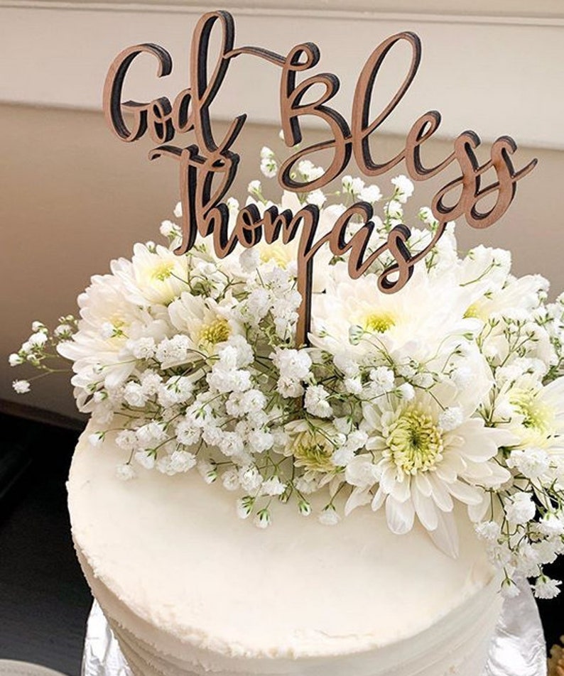 May include: A white cake with white flowers and a wooden cake topper that says "God Bless Thomas".