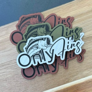 Puede incluir: Una colección de pegatinas marrones, verde oliva y blanquecinas con una ilustración de un pez lubina. Las pegatinas presentan el texto "Only Fins" en una fuente de escritura. Las pegatinas están dispuestas sobre una superficie de madera.