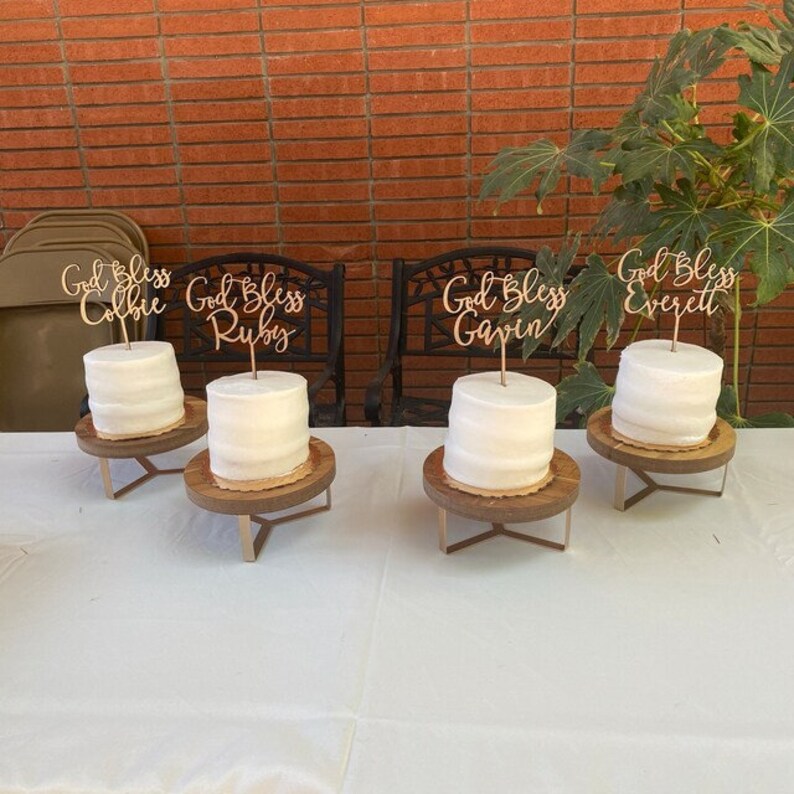 May include: Four white cakes with wooden cake stands and personalized wooden cake toppers that say "God Bless Colbie", "God Bless Ruby", "God Bless Gavin", and "God Bless Everett".