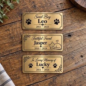 Op de afbeelding: Drie rechthoekige gedenkplaten van goudkleurig metaal met zwarte gravures. De bovenste plaat toont Sweet Boy Leo 2011-2023 met pootafdrukken. De middelste toont Faithful Friend Jasper 2010-2024 met een kat. De onderste toont In Loving Memory of Lucky 2011-2024 met pootafdrukken.