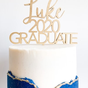 May include: A white cake with blue frosting and gold accents topped with a wooden cake topper that says "Luke 2020 Graduate".