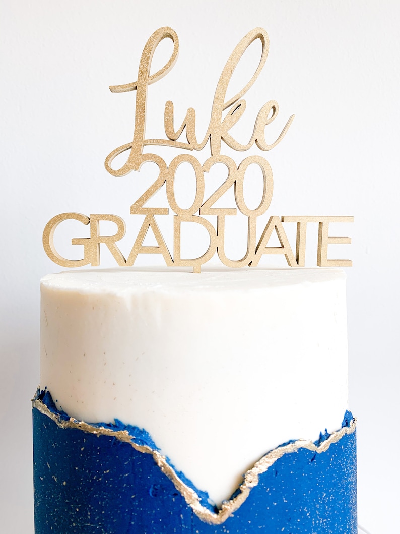 May include: A white cake with blue frosting and gold accents topped with a wooden cake topper that says "Luke 2020 Graduate".