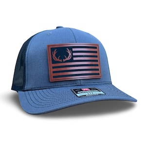 Könnte beinhalten: Eine graue und schwarze Trucker-Kappe mit einem braunen Leder-Patch, der eine amerikanische Flagge mit einem Hirschgeweih-Design zeigt. Der Patch trägt den Text "Richardson 112" unten.