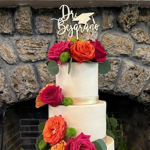 May include: A white wedding cake decorated with red, orange, and green flowers. The cake has a gold ribbon around the bottom tier and a wooden cake topper that says "Dr. Begrano".