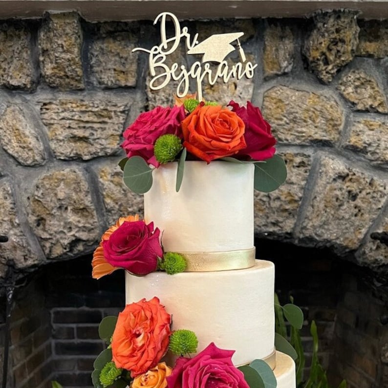 May include: A white wedding cake decorated with red, orange, and green flowers. The cake has a gold ribbon around the bottom tier and a wooden cake topper that says "Dr. Begrano".