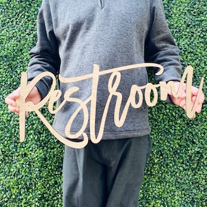 May include: A wooden sign with the word "Restroom" written in cursive script. The sign is held by a person in front of a green wall.
