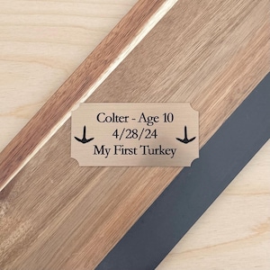 May include: A wooden plaque with a decorative border and a brass nameplate. The nameplate reads "Colter - Age 10, 4/28/24, My First Turkey" with arrow symbols. The plaque has a diagonal design with wood and dark gray accents.