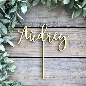 May include: A wooden cake topper with the name "Audrey" written in cursive script. The topper is gold in color and is attached to a thin wooden stick.