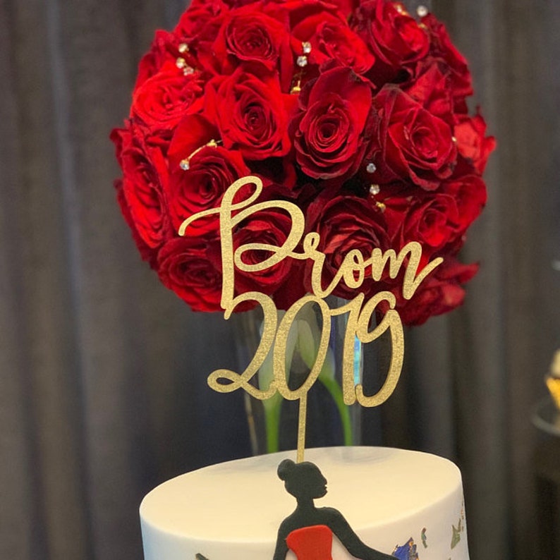 May include: Gold glitter cake topper with the words "Prom 2019" in a cursive font. The topper is placed on top of a white cake with a red rose bouquet in the background.