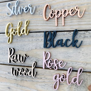 K&ouml;nnte beinhalten: Nahaufnahme von sechs Holzschildern mit verschiedenen Oberfl&auml;chen. Die Schilder zeigen "Silber", "Kupfer", "Gold", "Schwarz", "Rohholz" und "Ros&eacute;gold".