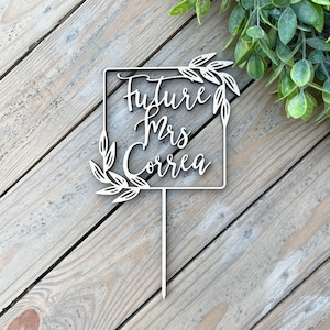 Puede incluir: Topper de pastel de madera con un marco cuadrado y acentos de hojas. El texto "Future Mrs. Correa" está escrito en cursiva dentro del marco.