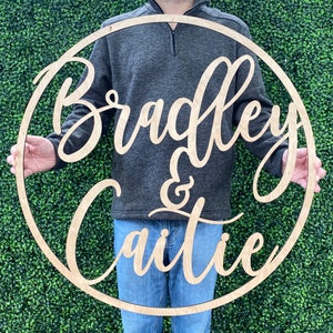 May include: A wooden circular sign with the names "Bradley & Caitie" in a flowing script font. The names are cut out of the light-colored wood, set against a dark background. The sign is held by a person.