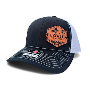 Może przedstawiać: Czarna i biała czapka trucker z brązową skórzaną naszywką z napisem "Florida - Est. 1845" z wzorem palmy i sylwetki dzikich zwierząt.