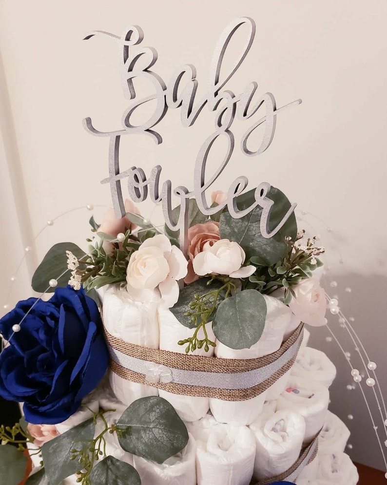 May include: A diaper cake decorated with blue and pink flowers, greenery, and a wooden sign that says "Baby Shower".